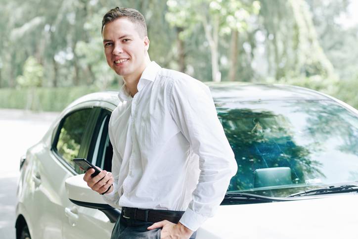 Porträt eines jungen Mannes, der ein Mobiltelefon in der Hand hält, in der Nähe seines Autos steht und in die Kamera lächelt
